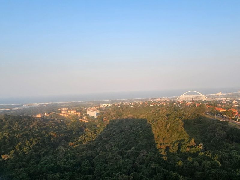 An elevated panoramic view overlooking a lush green landscape, urban residential areas, and the distant coastline.