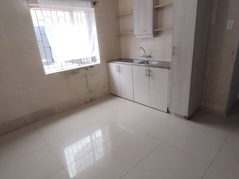 A compact, empty kitchen featuring tiled walls and floors, built-in cabinetry, and a stainless steel sink, showing signs of moisture damage on the wall.