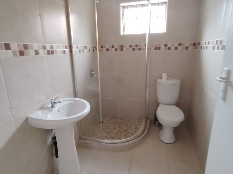 A clean, functional bathroom featuring a corner shower enclosure, pedestal basin, and toilet with neutral wall and floor tiling.