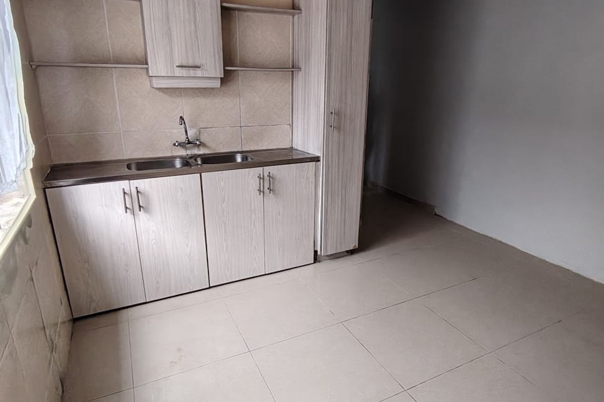 A compact kitchen area featuring light wood-grain cabinetry, a stainless steel sink, tiled backsplash, and neutral floor tiles in an empty, unfinished space.