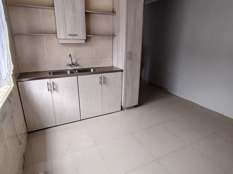 A compact kitchen area featuring light wood-grain cabinetry, a stainless steel sink, tiled backsplash, and neutral floor tiles in an empty, unfinished space.