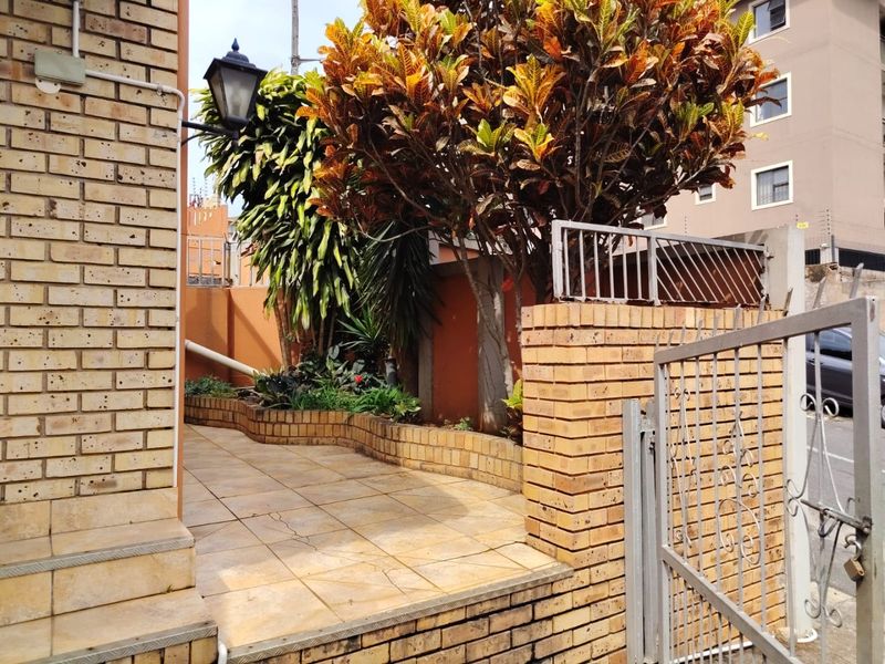 Front entrance area featuring a brick boundary wall, metal gate, tiled patio, and built-in brick planter with established landscaping.