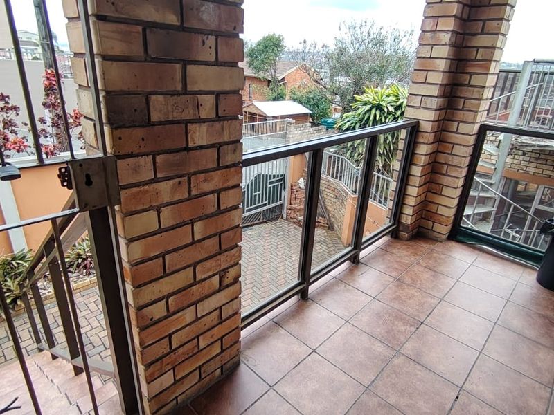 A covered balcony featuring brick pillars, tiled flooring, and a glass balustrade, offering an elevated view of the property's exterior and driveway area.
