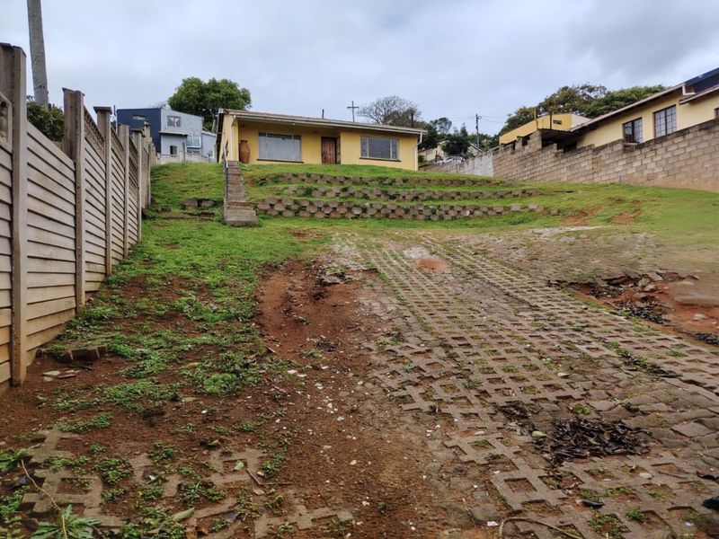 Front view of a single-story house featuring a sloped yard, concrete retaining walls, and a paved driveway requiring maintenance.