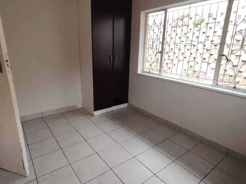 A compact, empty bedroom featuring tiled flooring, a built-in cupboard, and a window fitted with security bars.