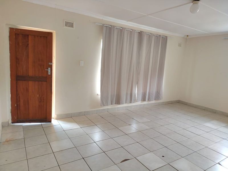 An empty living room featuring neutral tiled flooring, a solid wooden door, and simple ceiling lighting, offering a blank canvas for interior design.
