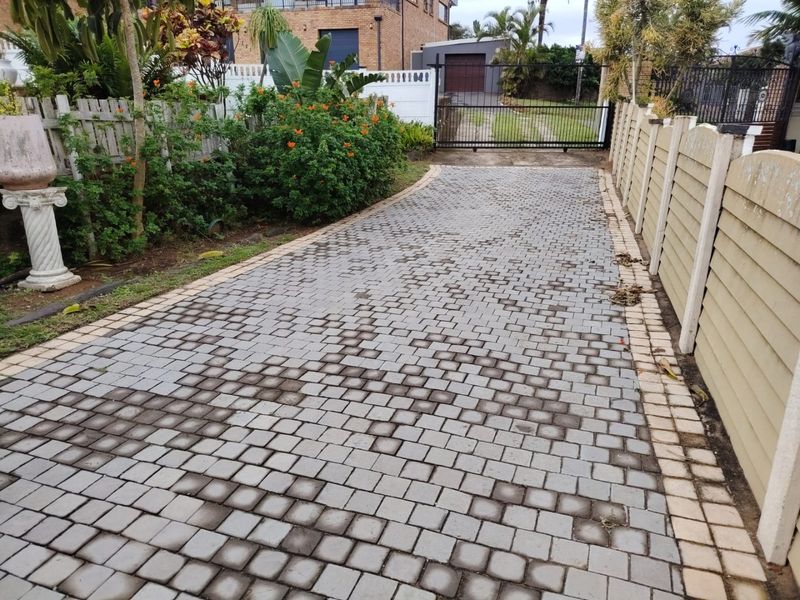 A paved driveway leading to a metal security gate, bordered by a precast concrete wall and established garden beds.