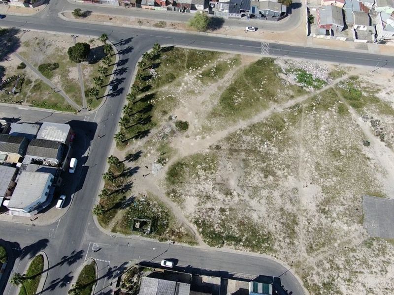 An aerial view of a vacant residential plot situated at a street corner, featuring natural vegetation and surrounding road infrastructure.