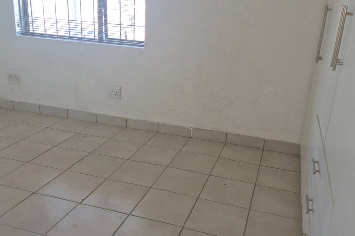 An empty bedroom featuring neutral tiled flooring, built-in white cupboards, and a window fitted with security bars.