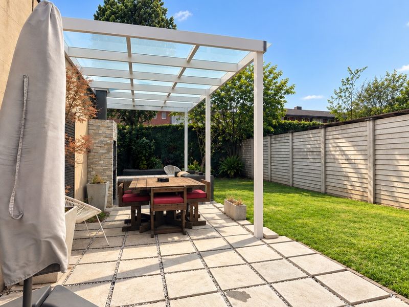 A well-maintained outdoor patio featuring a modern glass-roofed pergola, built-in fireplace, stone paving, and a private garden area with timber fencing.
