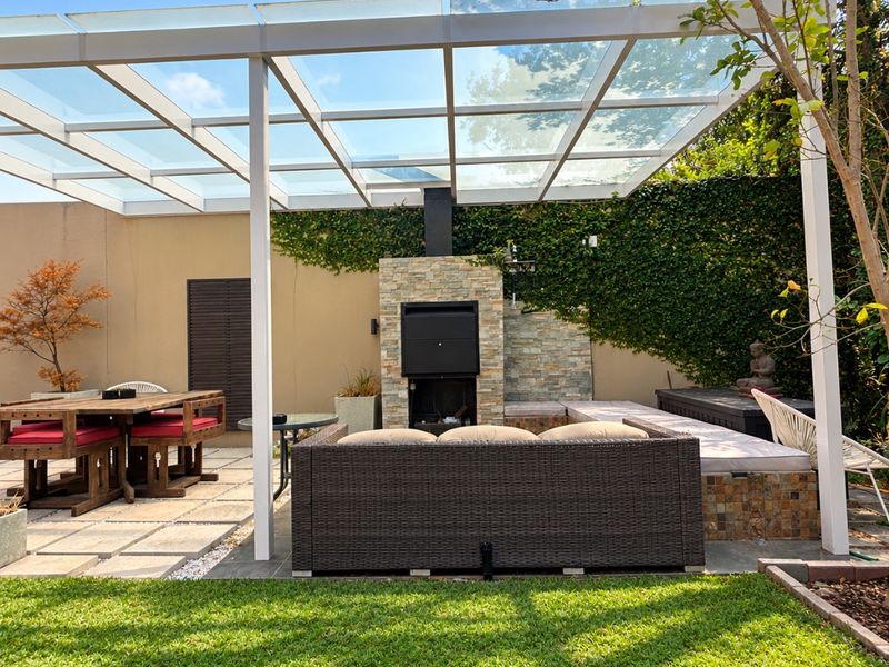A well-maintained outdoor patio featuring a modern glass-roofed pergola, built-in braai with stone cladding, and integrated seating area overlooking the garden.