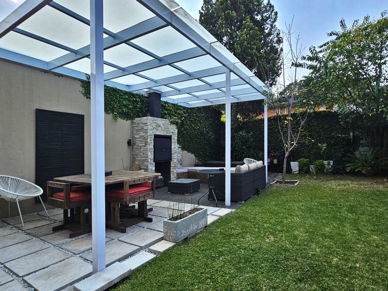 A well-maintained outdoor patio featuring a modern white pergola, built-in stone fireplace, and paved flooring, overlooking a private garden area.