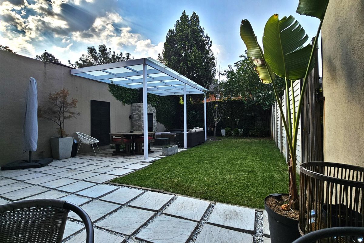 A well-maintained rear garden featuring a paved patio area with a modern pergola and a built-in outdoor fireplace, complemented by a neat lawn.