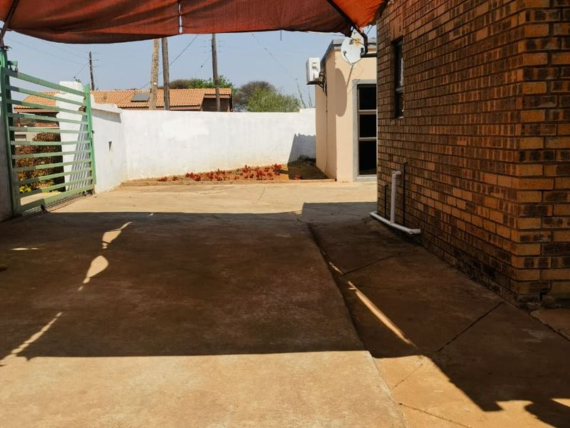 A paved rear exterior area featuring a brick wall, metal gate, and a large overhead shade sail providing coverage for the outdoor space.