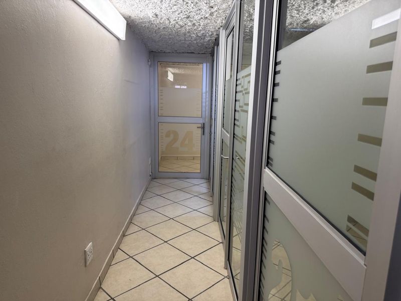 A commercial hallway featuring tiled flooring, textured ceilings, and glass partition walls with frosted privacy panels leading to an exterior door.
