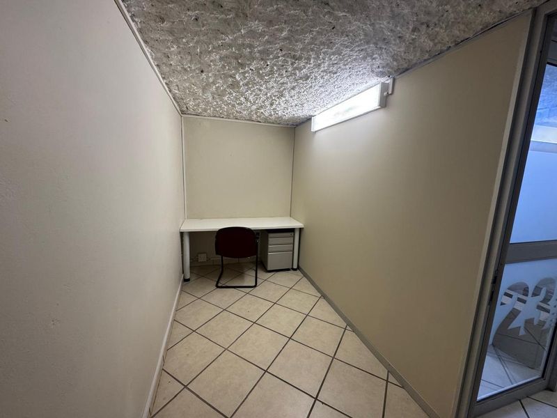 A compact office space featuring tiled flooring, a textured ceiling, and a fluorescent light fixture, providing a functional work environment.