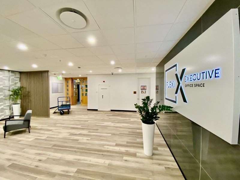 Modern commercial office reception area featuring wood-look flooring, a suspended ceiling with recessed lighting, and a prominent illuminated company signage wall.