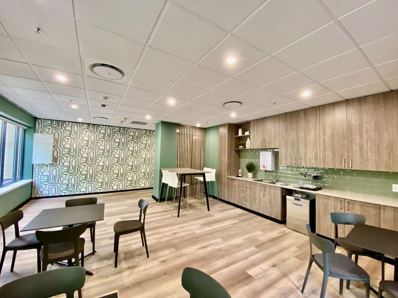 A modern commercial kitchenette featuring wood-look cabinetry, integrated appliances, tiled backsplash, and a suspended ceiling with recessed lighting in an open-plan office setting.