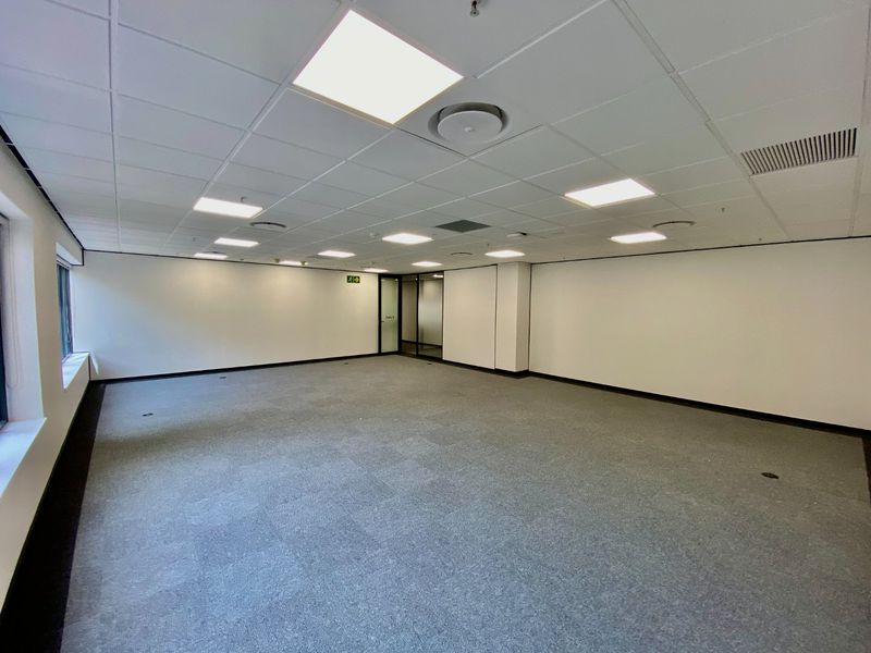 A spacious, empty commercial office suite featuring a suspended ceiling with recessed lighting, carpeted flooring, and a glass entrance door.