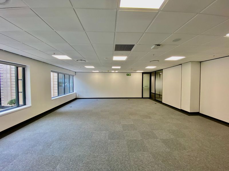 A spacious, vacant commercial office suite featuring neutral carpet tiles, a suspended ceiling with recessed lighting, and large windows providing natural light.