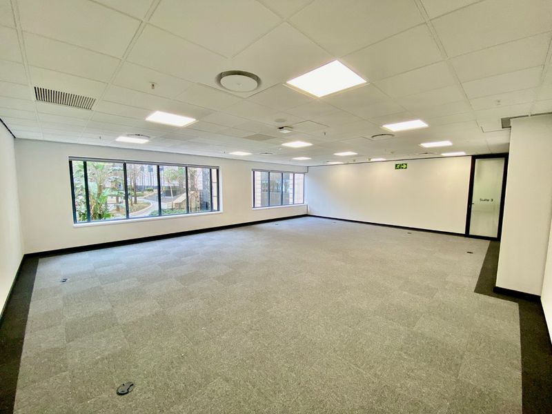 A spacious, vacant commercial office suite featuring neutral carpet tiles, a suspended ceiling with recessed lighting, and large windows providing natural light.