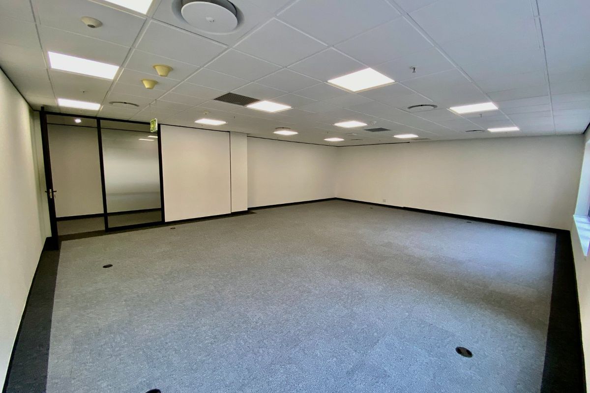 A spacious, empty commercial office area featuring a suspended ceiling with recessed lighting, carpeted flooring, and a modern glass partition wall.
