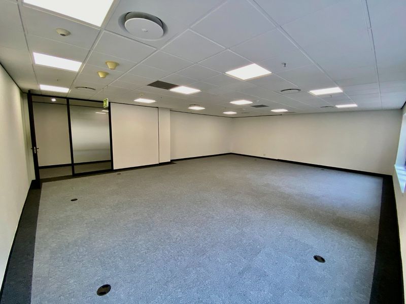 A spacious, empty commercial office area featuring a suspended ceiling with recessed lighting, carpeted flooring, and a modern glass partition wall.