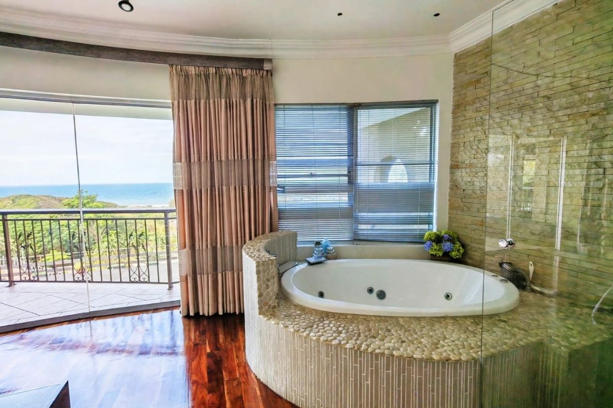 Luxurious bathroom featuring a built-in jetted tub with stone cladding, glass shower enclosure, hardwood floors, and direct access to a balcony with ocean views.