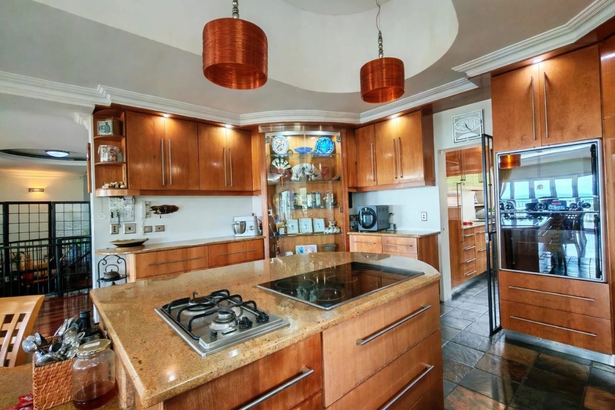 Spacious kitchen featuring warm wooden cabinetry, granite countertops, a central island with dual hobs, and a unique recessed ceiling dome with pendant lighting.