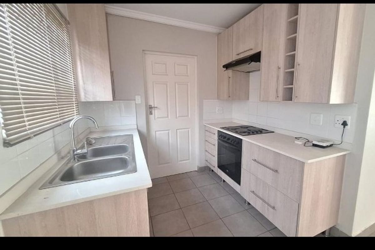 A functional kitchen featuring light wood-tone cabinetry, a double stainless steel sink, built-in oven with hob, extractor fan, and neutral tiled flooring.