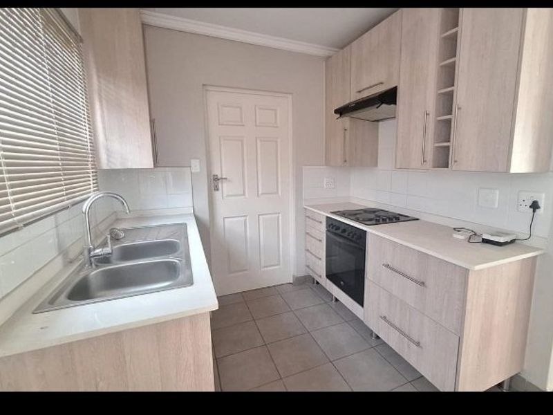A functional kitchen featuring light wood-tone cabinetry, a double stainless steel sink, built-in oven with hob, extractor fan, and neutral tiled flooring.