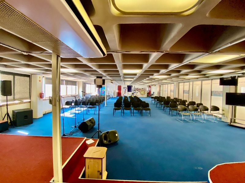 Spacious commercial event hall featuring a raised stage, distinctive coffered ceiling design, and large windows providing natural light.