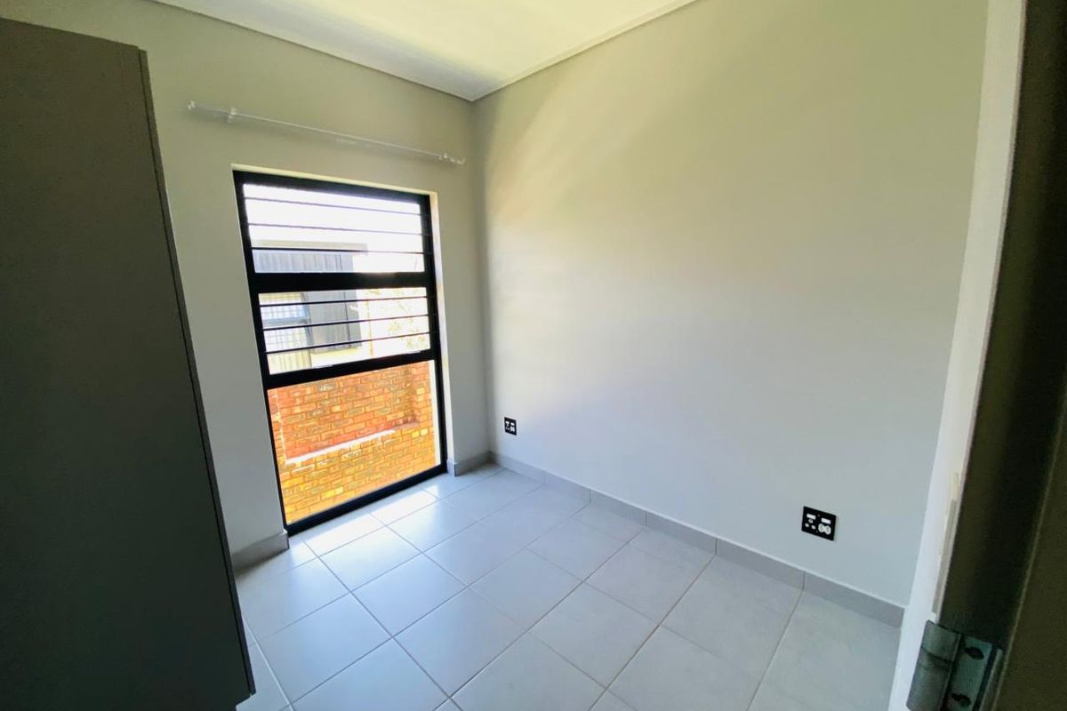A clean, empty bedroom featuring neutral tiled flooring, a built-in cupboard, and a window fitted with security bars.