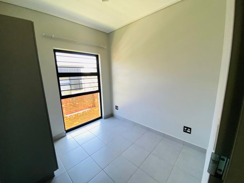 A clean, empty bedroom featuring neutral tiled flooring, a built-in cupboard, and a window fitted with security bars.