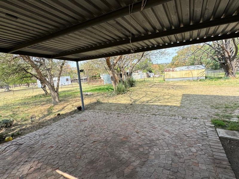 A covered outdoor patio area featuring a corrugated metal roof and cobblestone paving, overlooking a spacious garden area.
