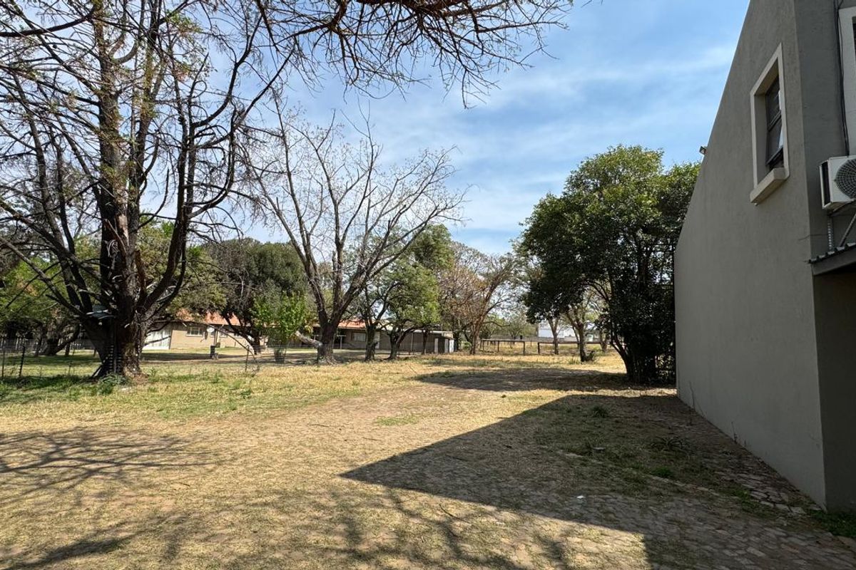 A view of the rear exterior area featuring a paved section, a wall-mounted air conditioning unit, and a spacious garden with mature trees.