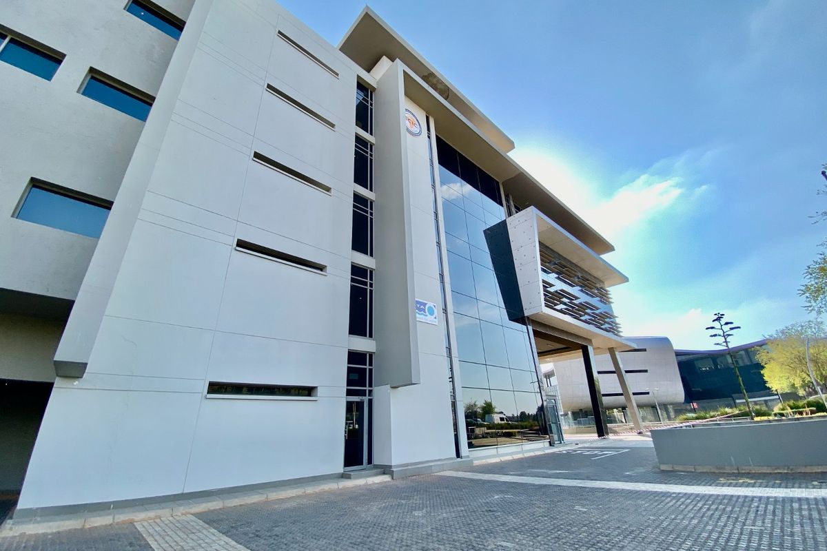 Modern commercial office building featuring a striking glass curtain wall facade, concrete structural elements, and a paved entrance area.