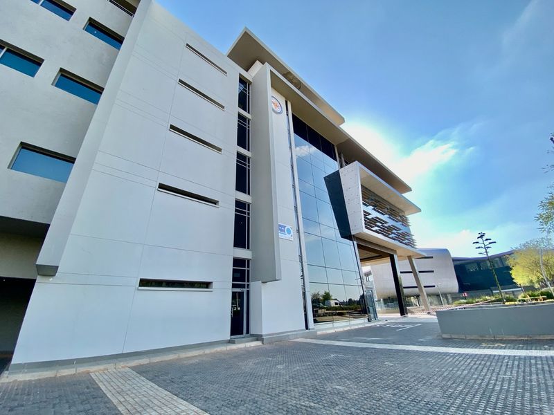 Modern commercial office building featuring a striking glass curtain wall facade, concrete structural elements, and a paved entrance area.