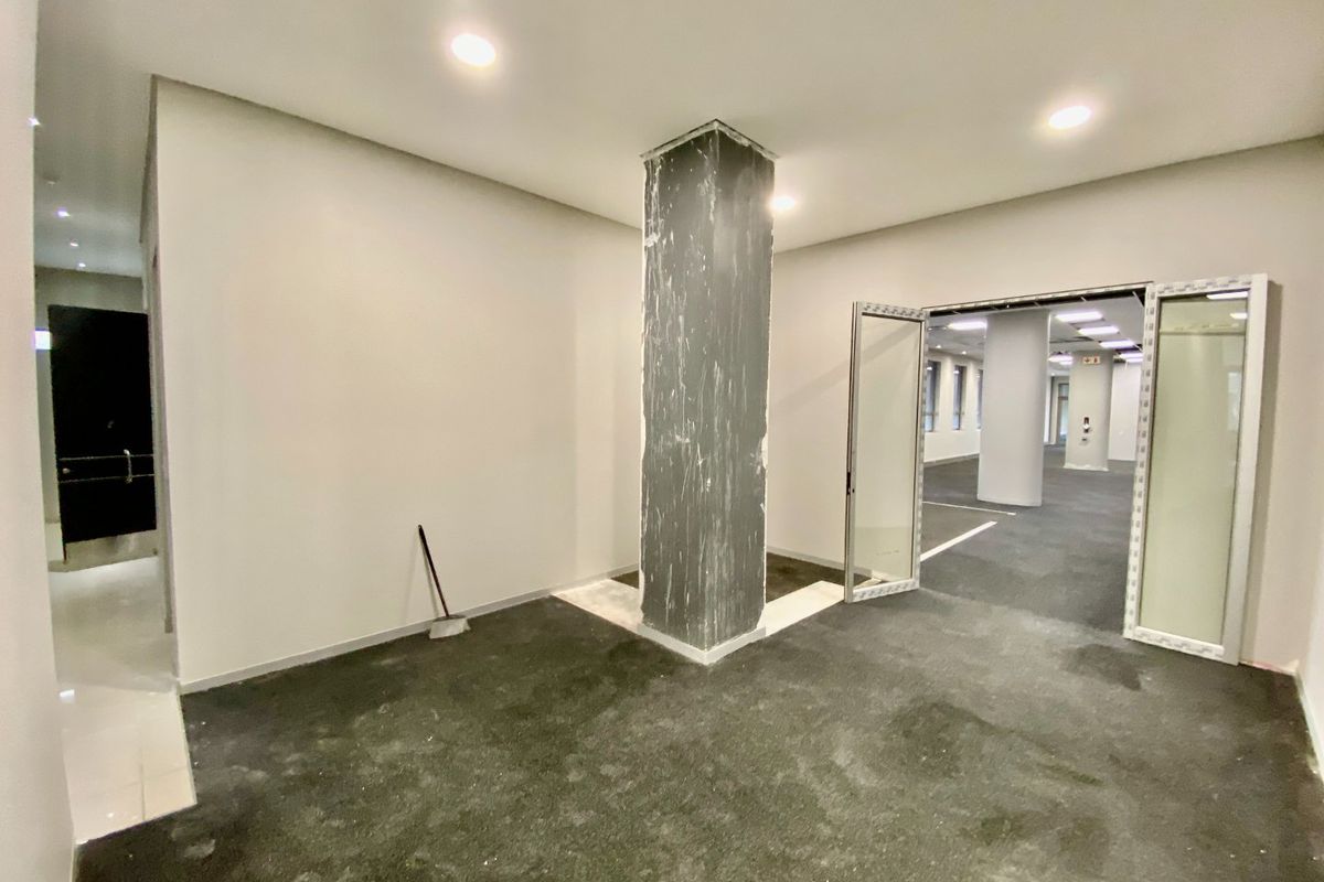 An empty commercial office space featuring a central structural column, recessed lighting, and double glass doors leading to an adjacent workspace.