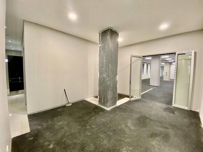 An empty commercial office space featuring a central structural column, recessed lighting, and double glass doors leading to an adjacent workspace.