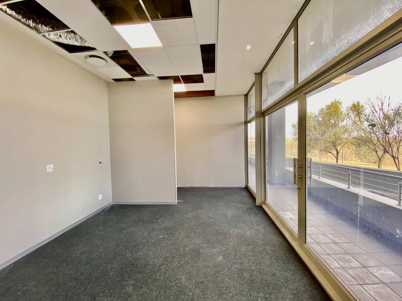 Empty commercial office space featuring large glass sliding doors leading to a balcony, carpeted flooring, and a suspended ceiling requiring maintenance.
