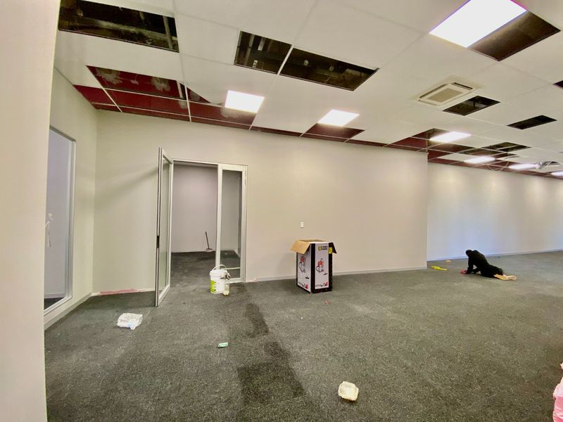 An open-plan commercial office space featuring carpeted flooring, glass partition doors, and a suspended ceiling currently undergoing maintenance with missing tiles.