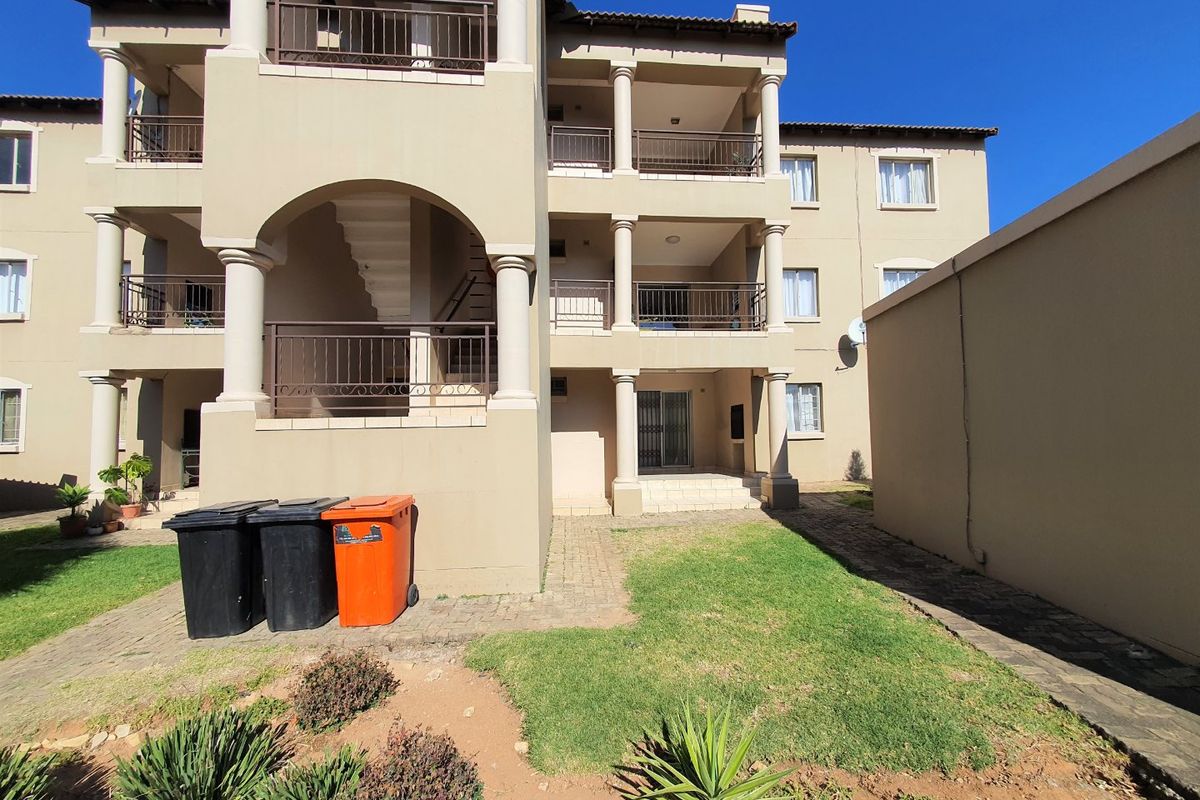 Rear view of a multi-story apartment complex featuring balconies with metal railings, paved walkways, and a small landscaped lawn area.
