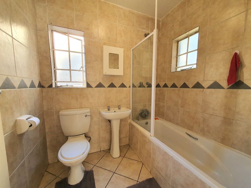 A dated bathroom featuring a pedestal basin, toilet, and a bathtub with a glass shower screen, finished with neutral wall and floor tiles.