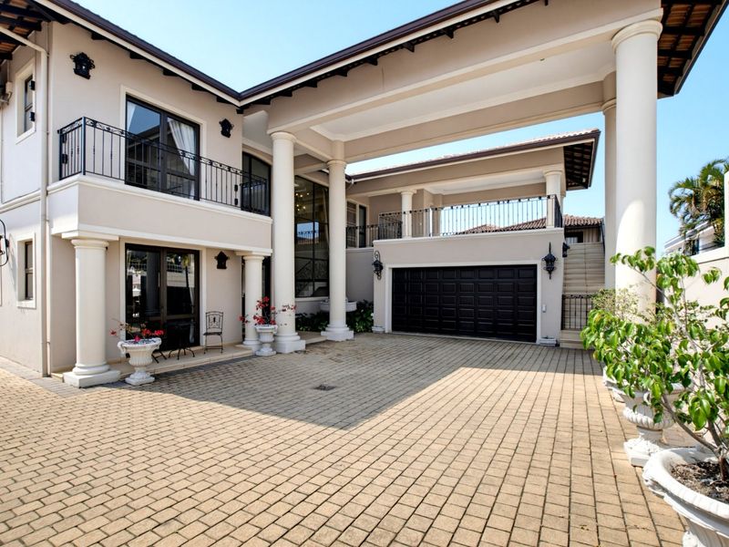 A well-maintained front exterior featuring a paved driveway, double garage, covered portico with classical columns, and an upper-level balcony with wrought iron railings.