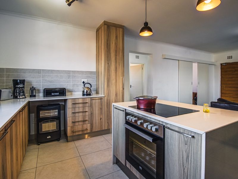 Modern kitchen featuring wood-grain cabinetry, stone countertops, a central island with a built-in oven and hob, and neutral tiled flooring.