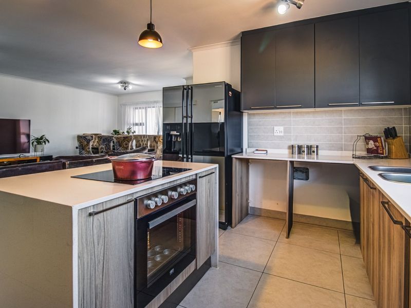 Modern open-plan kitchen featuring dark upper cabinetry, wood-grain lower cabinets, stone countertops, built-in oven, and hob, flowing into the adjacent living area.