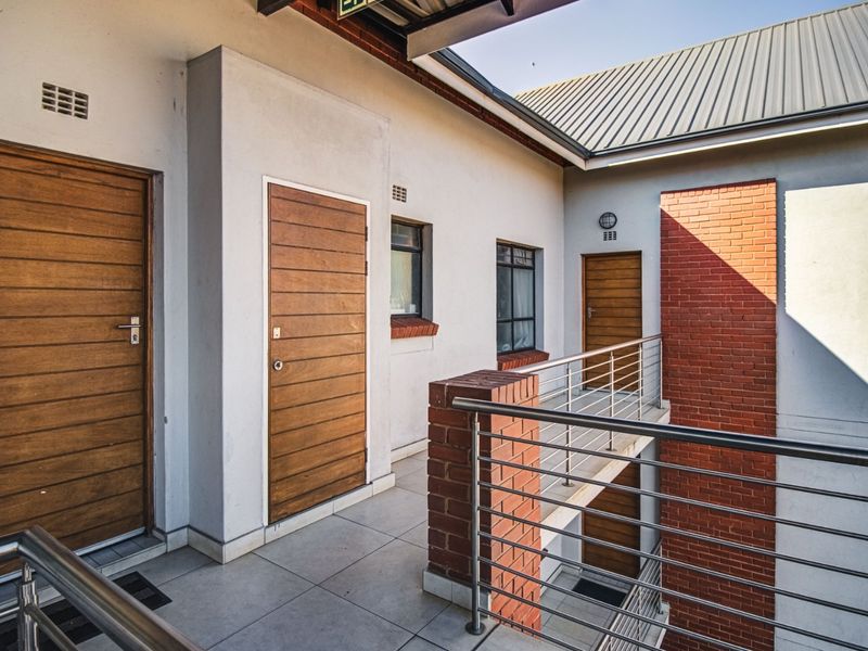 An exterior view of an apartment building balcony featuring tiled flooring, stainless steel railings, and brick accents with multiple timber entrance doors.