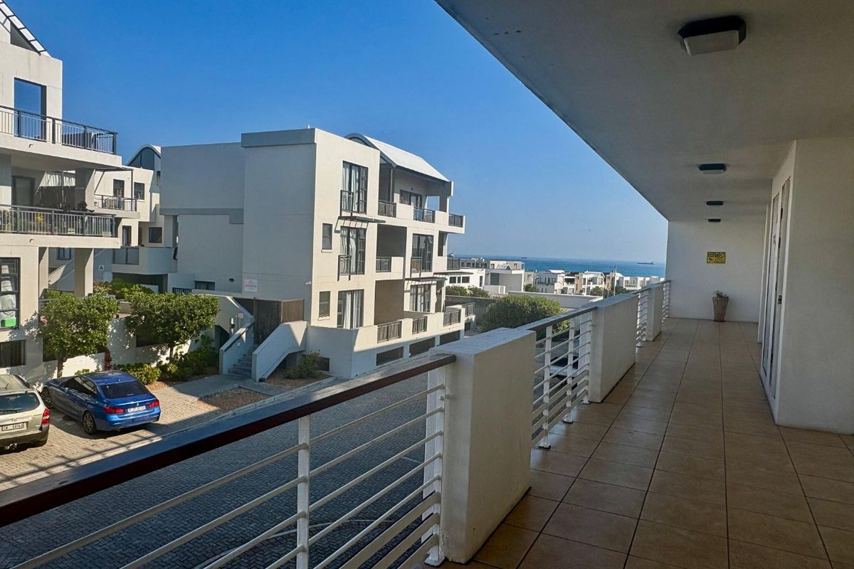A spacious covered balcony featuring tiled flooring and a metal balustrade, offering an elevated view of the surrounding residential complex and distant ocean.