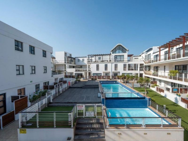 A well-maintained communal pool area featuring tiered swimming pools, timber decking, artificial grass, and modern metal balustrades within an apartment complex.